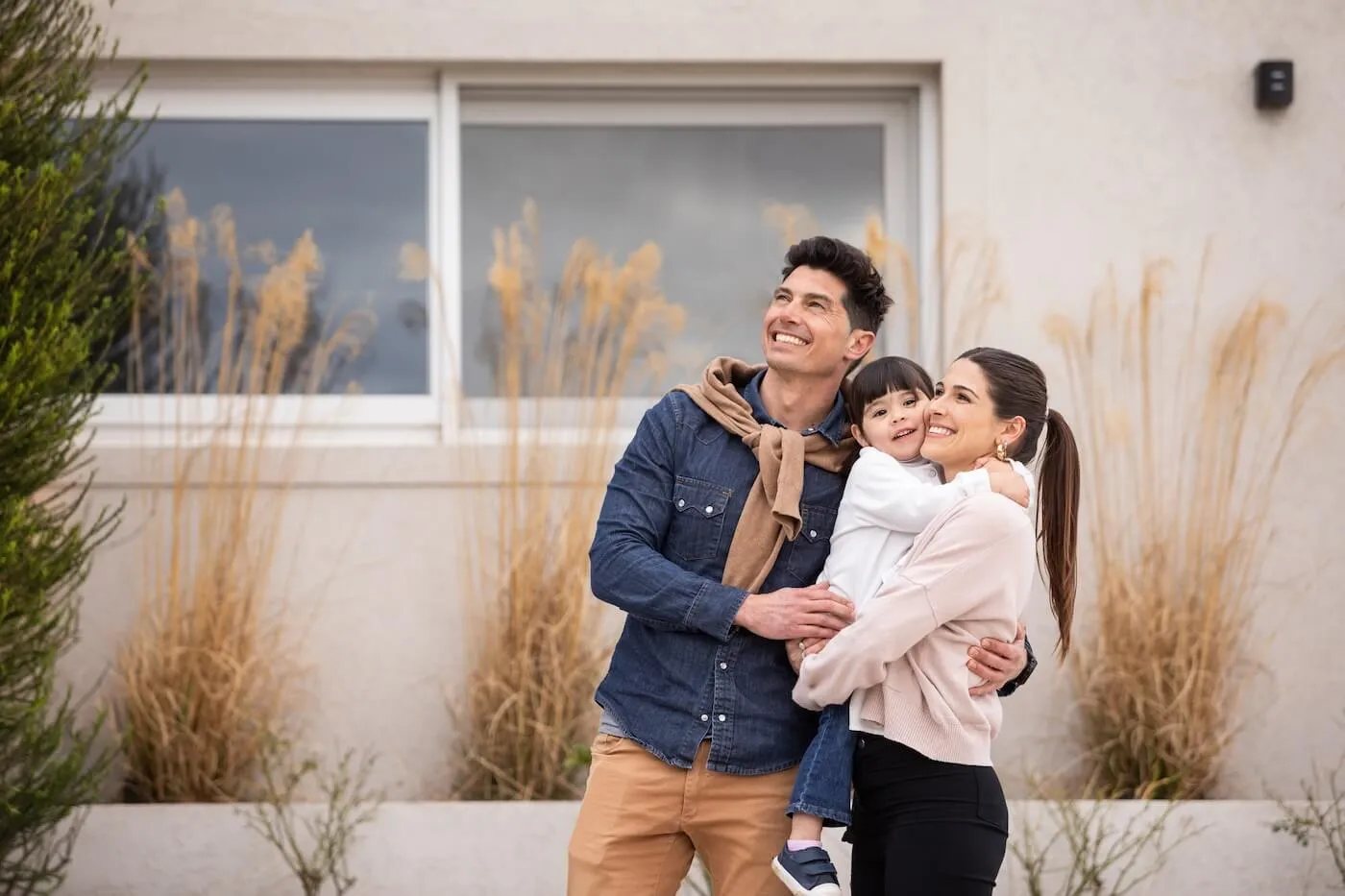 A couple stands outdoors holding a young child in their arms, posing together in front of their new house