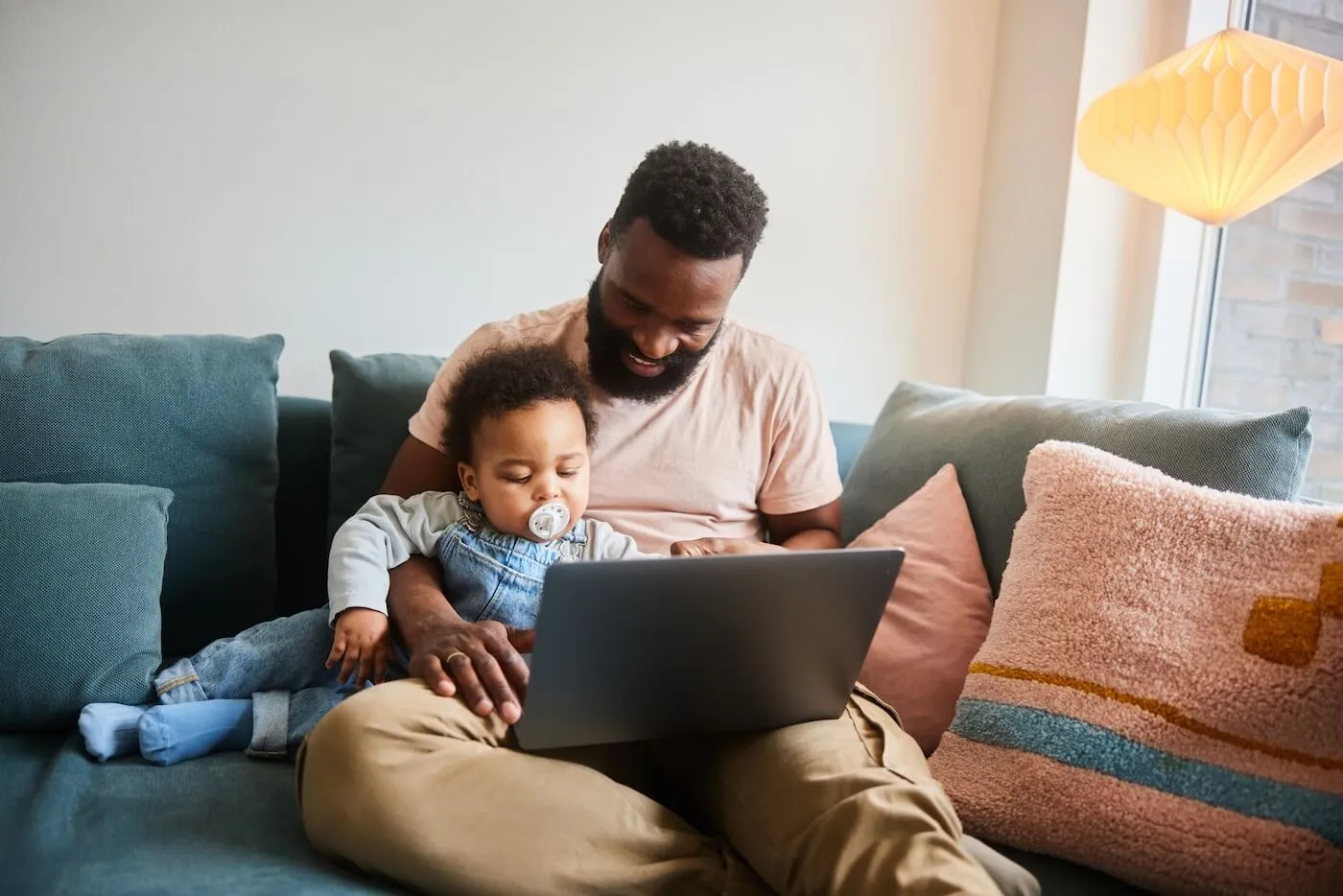 Smiling man sitting on a sofa with a baby on his lap, using a laptop together in a cozy living room. Decorative pillows, a floor lamp, and a large window are visible in the home setting.