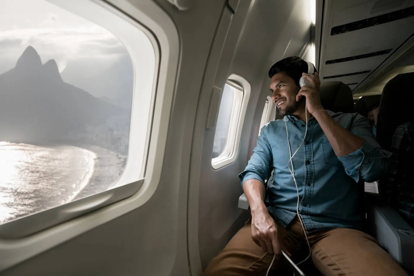 Airline passenger seated by an airplane window wearing headphones and looking out at a coastal landscape and mountains during a flight.