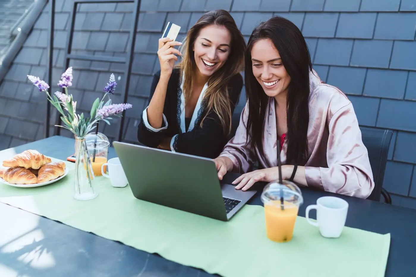 Two happy female friends sitting at an outdoor table with an open laptop, with one person holding a payment card; croissants, coffee cups, a glass of orange juice, and a small vase of flowers sit on the table, with a dark shingle wall and patio setting visible in the background.