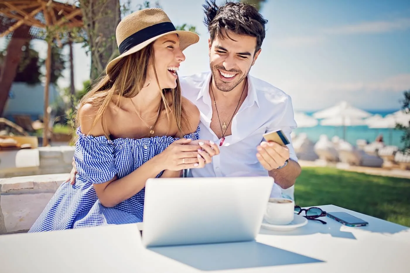 Happy couple seated at an outdoor table near a beach resort using a laptop together, with the man holding a payment card; a coffee cup, sunglasses, and a smartphone rest on the table, while lounge chairs, umbrellas, greenery, and the ocean are visible in the background under bright daylight.