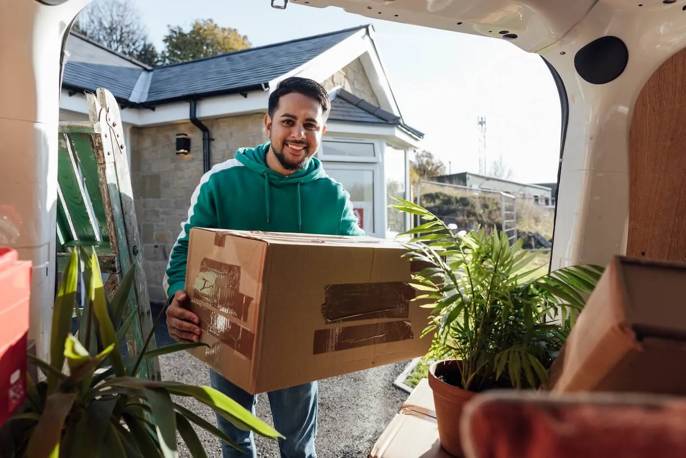 Smiling young man taking a moving box from the trunk of a car filled with boxes and potted plants