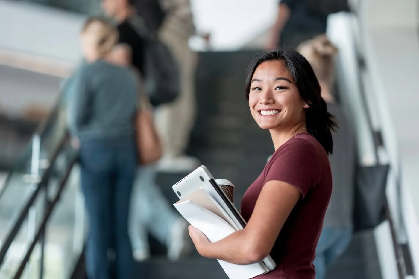 Smiling young woman holding printed documents, a laptop, and a paper cup, with a group of young people standing on stairs in the background