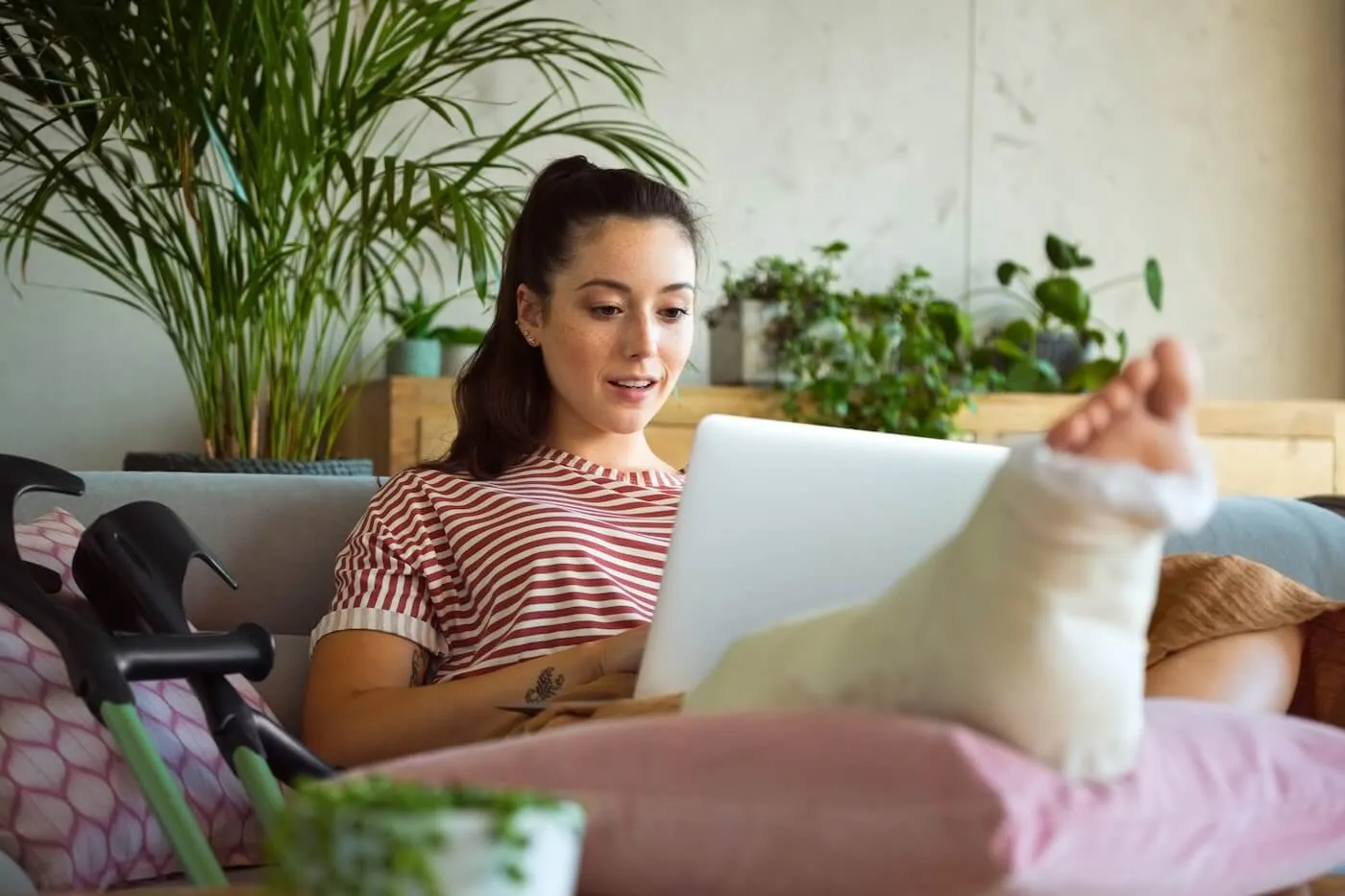 Young woman resting on a couch with her leg in a cast propped up on pillows, using a laptop, with crutches beside her