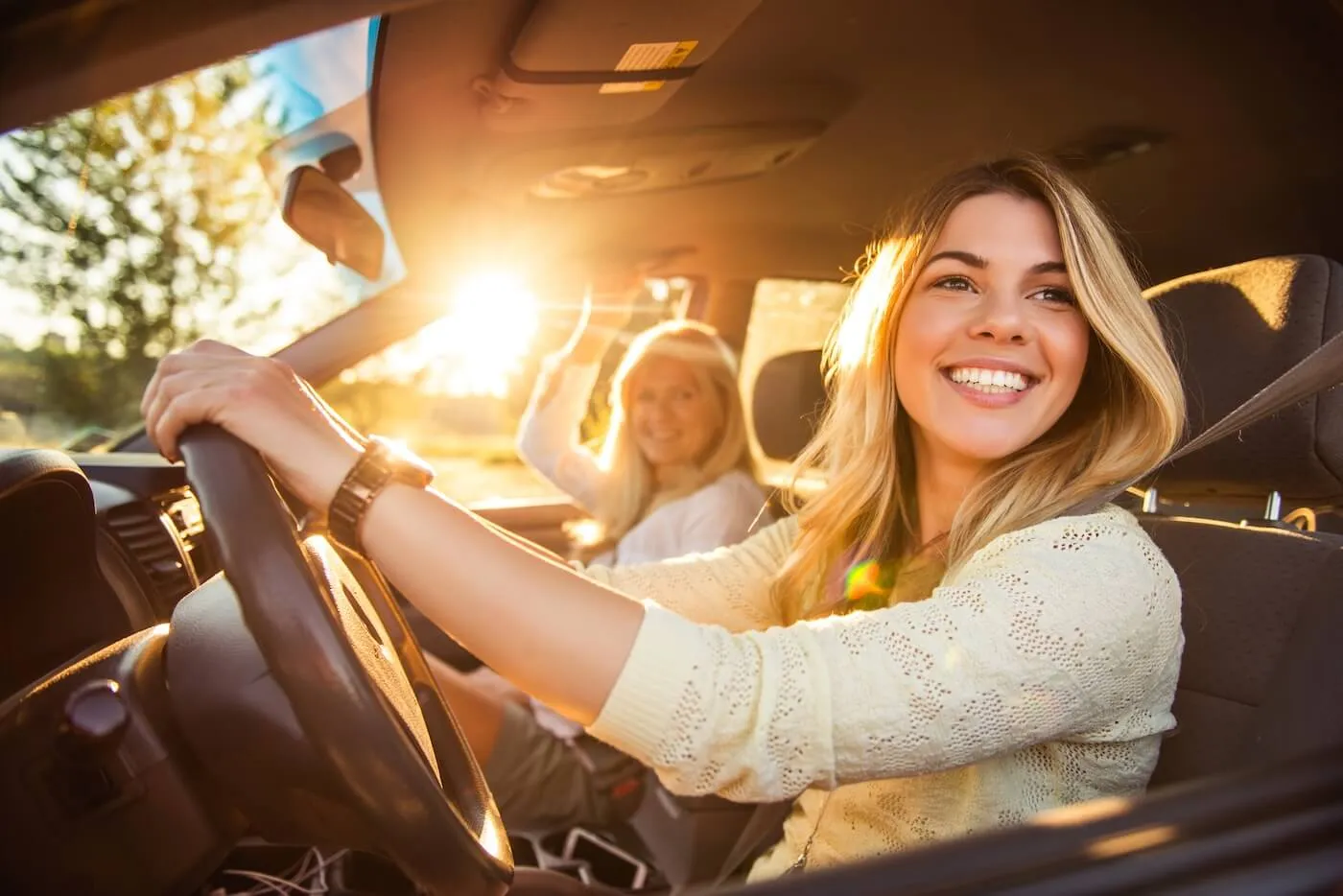 A cheerful young woman drives through the sunset, with a mature woman seated beside her in the passenger seat.