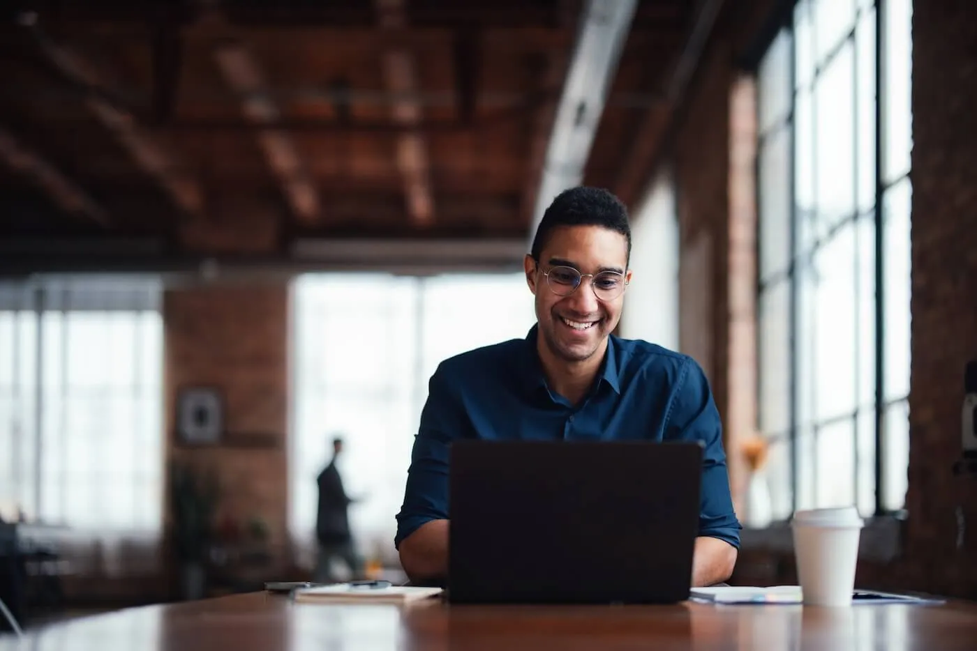 Smiling man using his laptop in a modern office open space, with a paper cup beside him