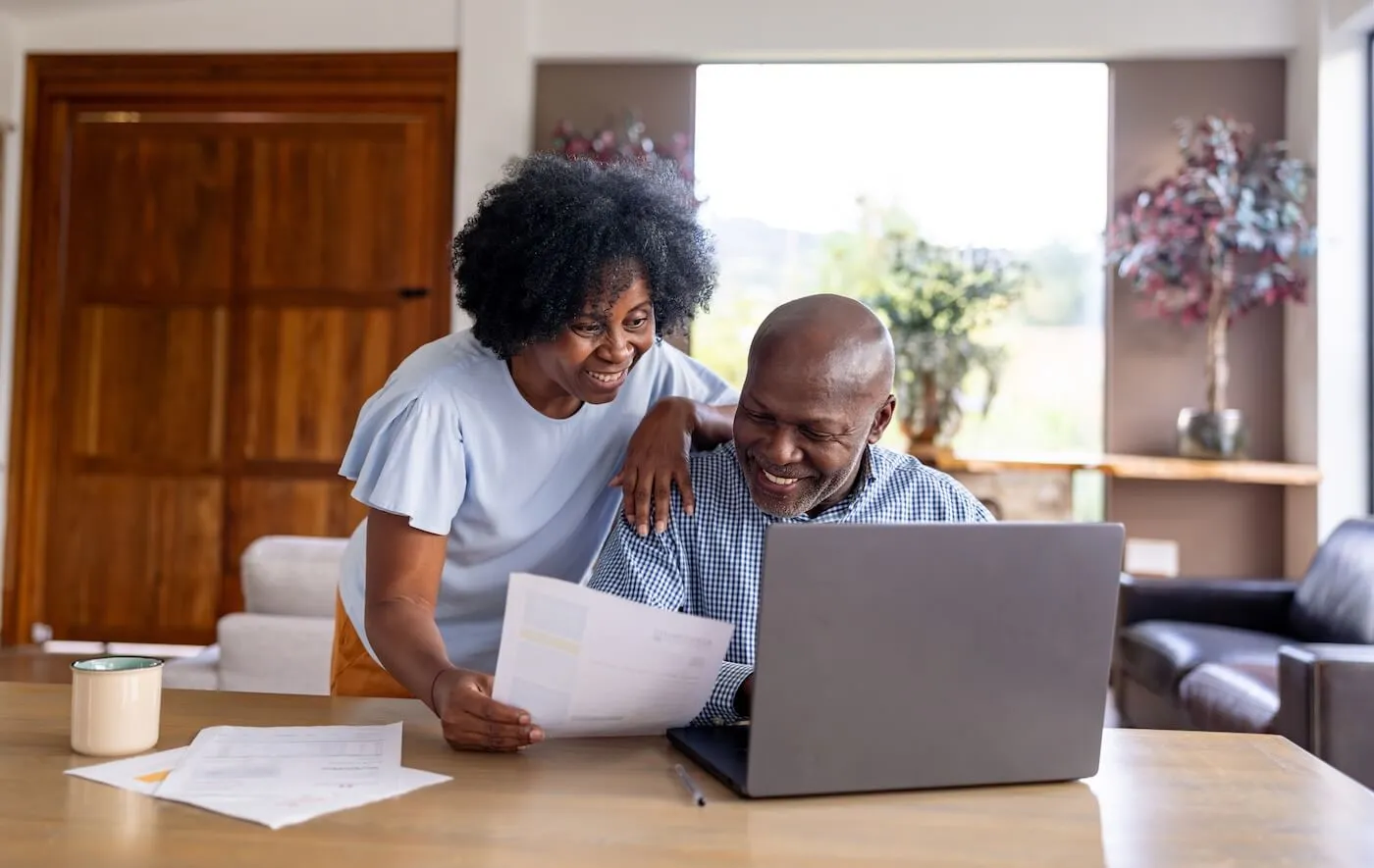 Happy couple sits at a table reviewing a document together, with a laptop open and papers spread out in a bright living room.