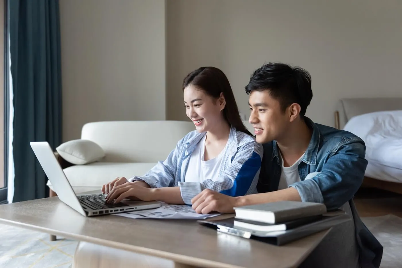 Young couple sitting at a low table at home, working together on a laptop with documents and books nearby.