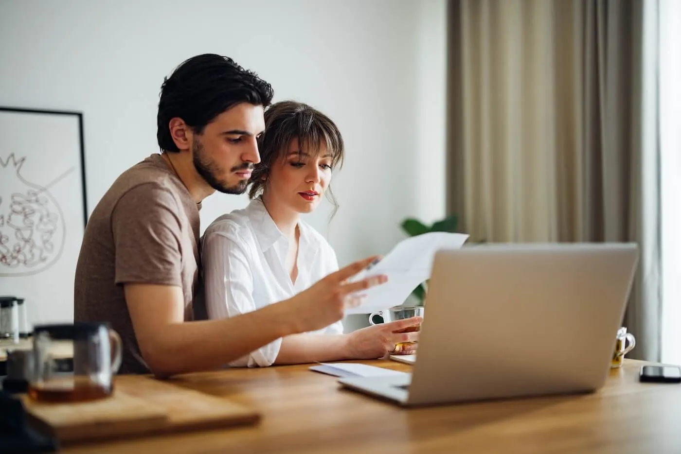 Focused couple reading their rental agreement while sitting at the desk with an open laptop, a mug, and a teapot