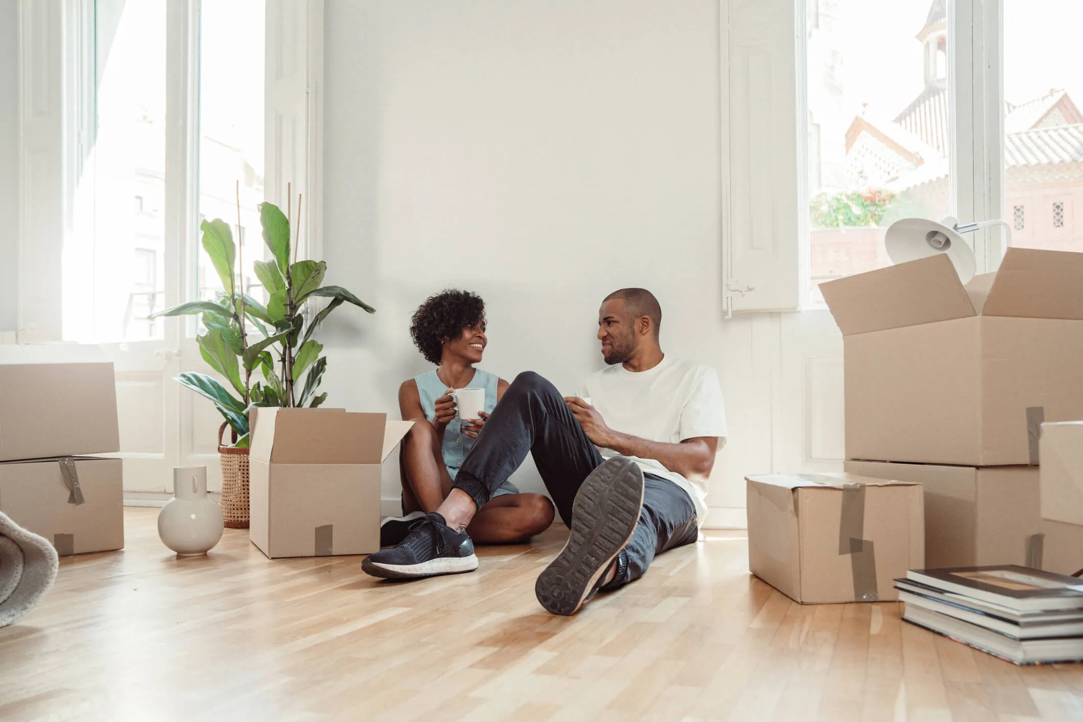 Relaxed couple sits on a wooden floor inside a bright room with cardboard boxes around them, each holding a mug, with a potted plant and stacked boxes nearby.