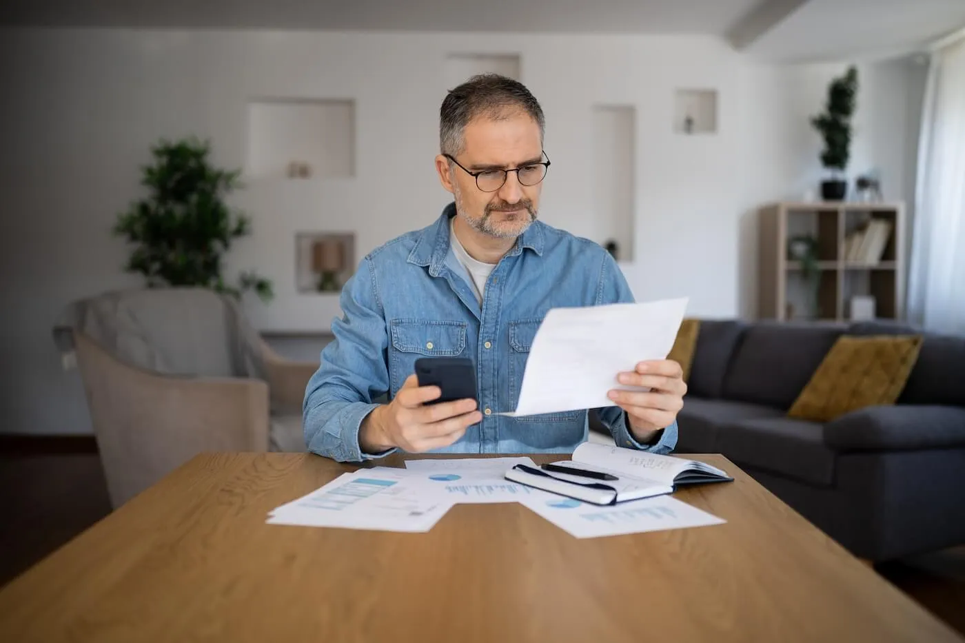Focused mature man reviewing a printed document while holding a smartphone, with printouts and notes on the table beside him