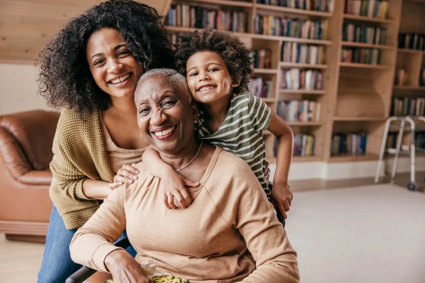 Three people posing for a photo, with a younger woman and a child standing behind and leaning toward the senior woman seated in front, with a large bookcase in the background.