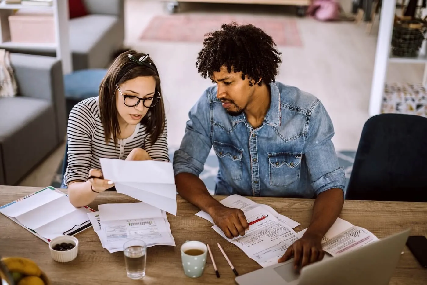 Concerned couple sitting at a wooden table, reviewing printed documents together, with a laptop open among scattered papers, pens, and cups in a home setting.