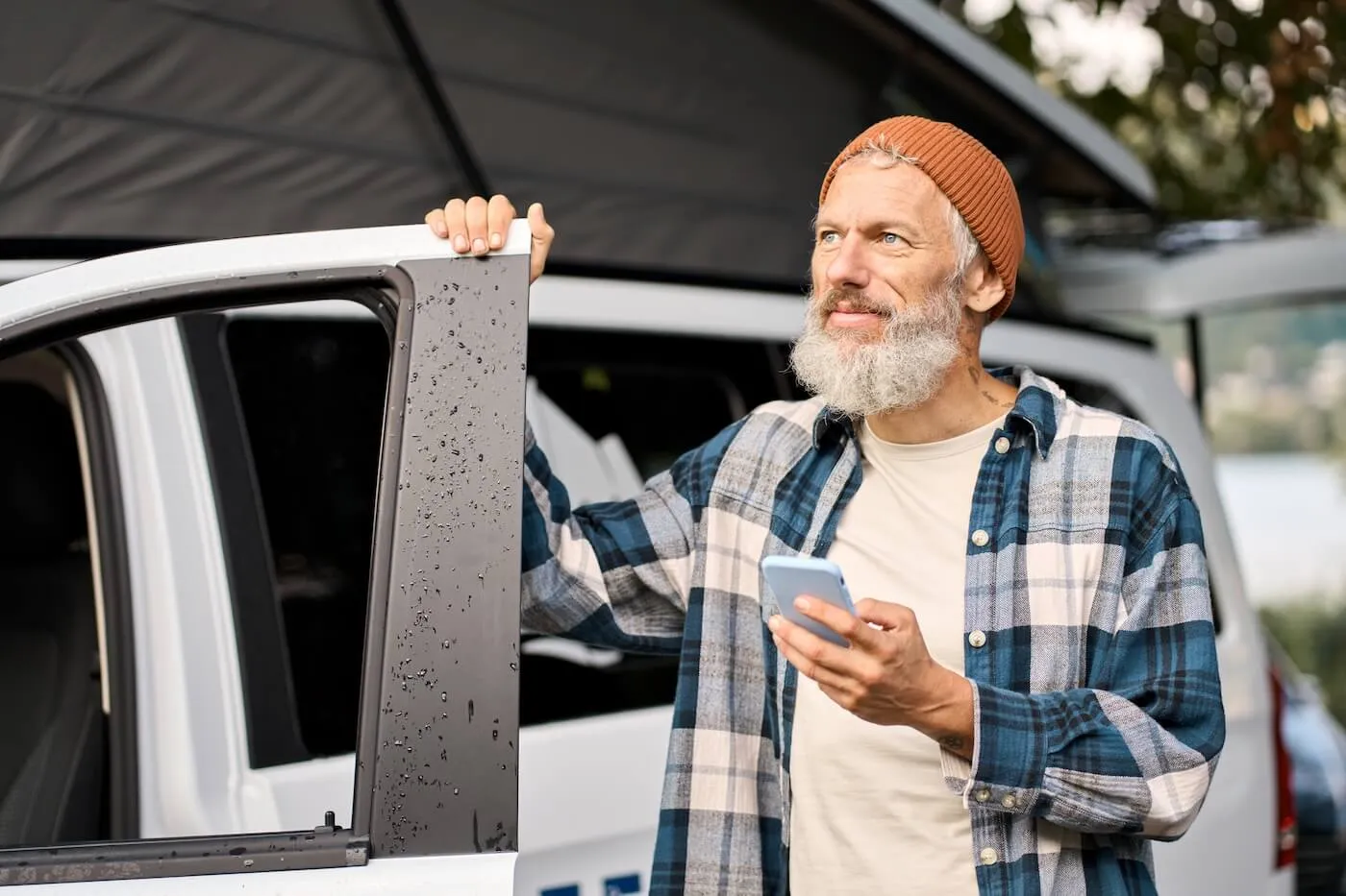 Mature man standing beside an open vehicle door while holding a smartphone, wearing a plaid shirt and beanie outdoors
