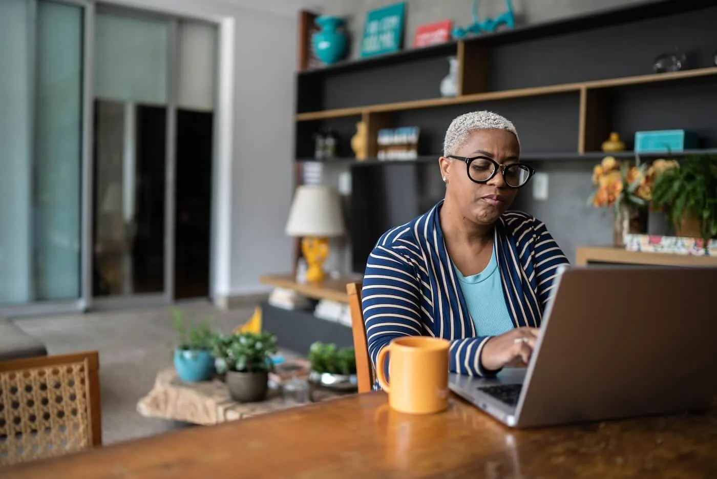 Focused mature woman wearing glasses is using her laptop at home, with a yellow mug beside her