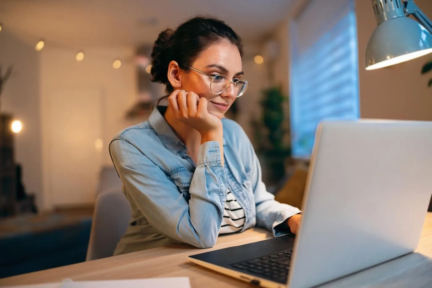 Pensive young woman sitting at a desk using a laptop, resting one hand on the face, with papers on the table and a desk lamp illuminating a home workspace in the background