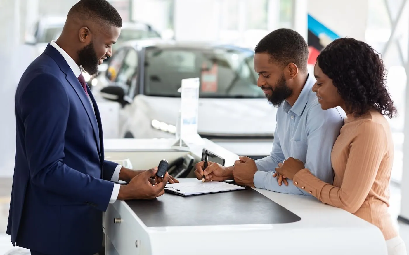 Car salesman meeting with a couple at a dealership counter, reviewing documents and discussing a vehicle purchase while holding car keys