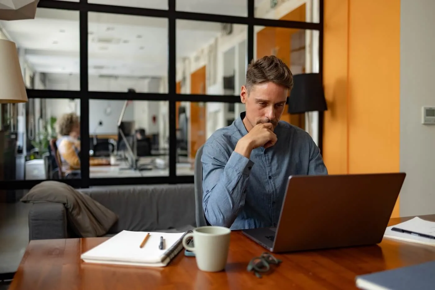 Focused man working on a laptop at a desk in a modern office, with notebooks, a pen, and a coffee mug on the table