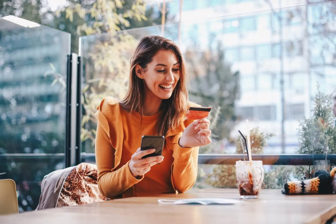 Smiling woman sits at an outdoor café table holding a credit card and using a smartphone, with a drink and personal items on the table and buildings in the background.