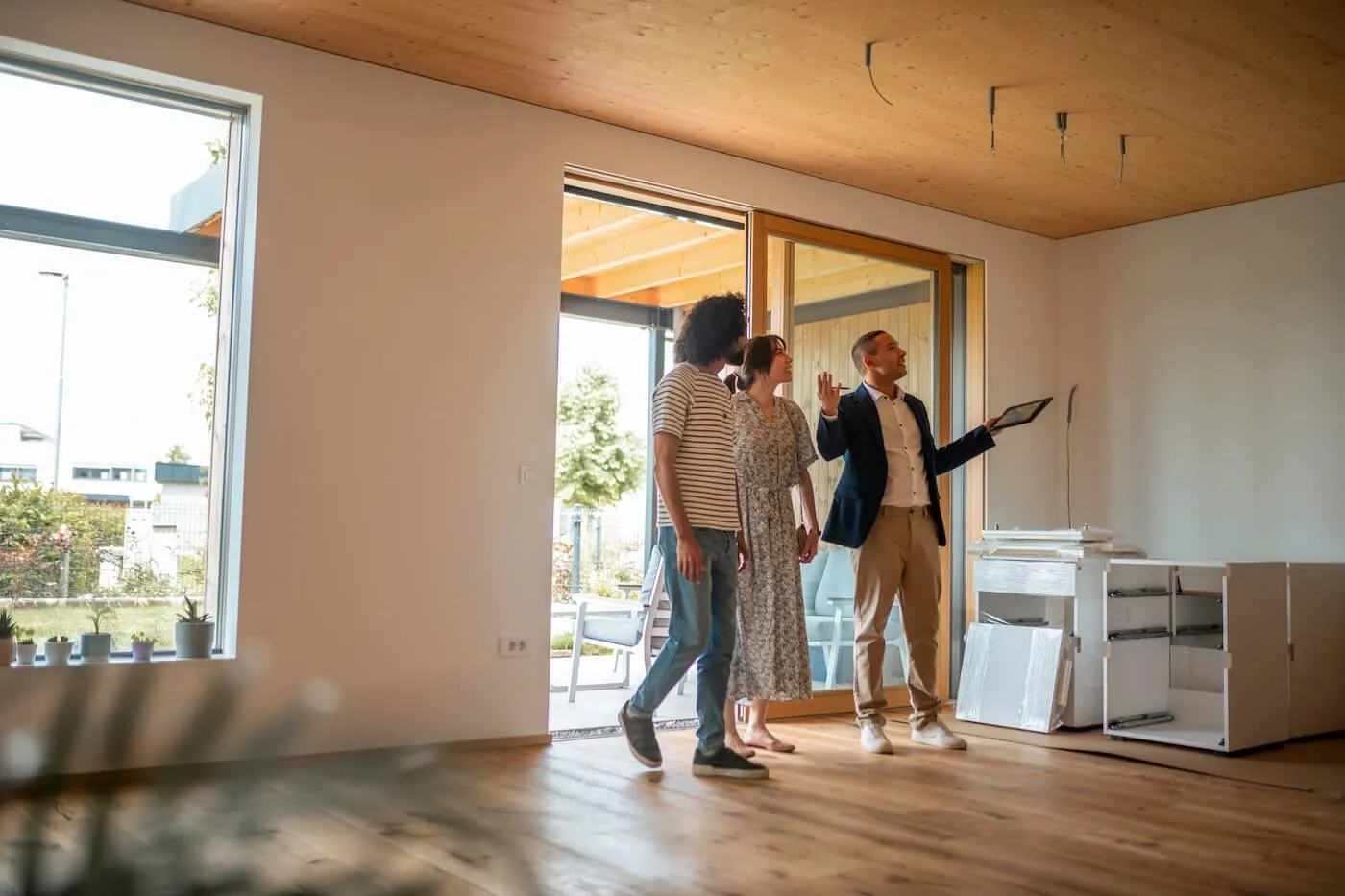 The couple stands inside an empty room while a real estate agent gestures toward the space with a tablet, with unpacked cabinetry nearby.