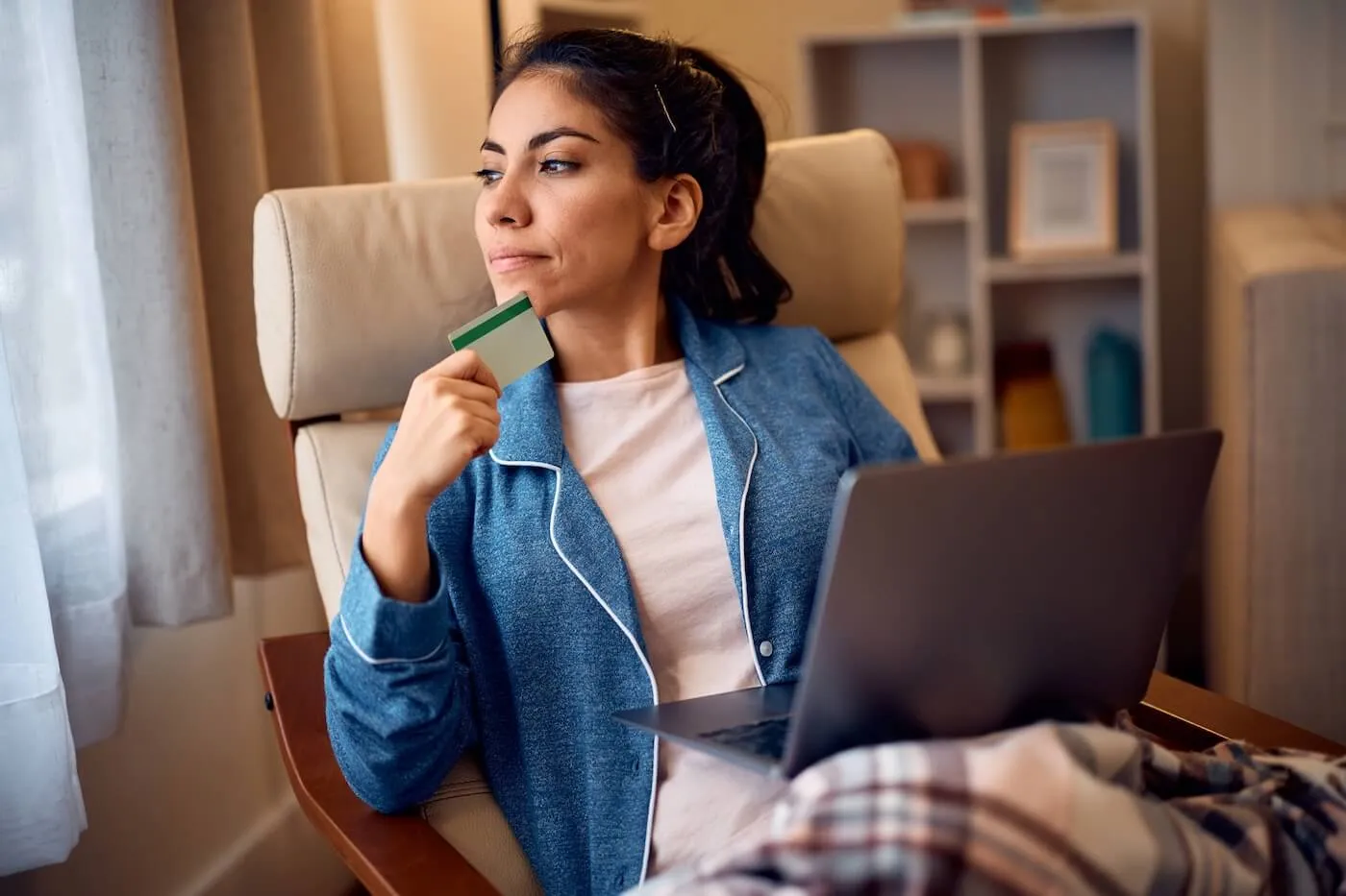 Pensive woman sitting in an armchair under a plaid blanket, holding a laptop and a credit card