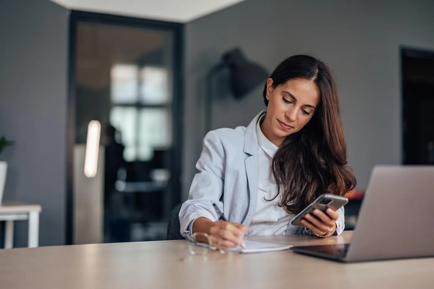 Focused woman making notes while using a laptop and a mobile phone