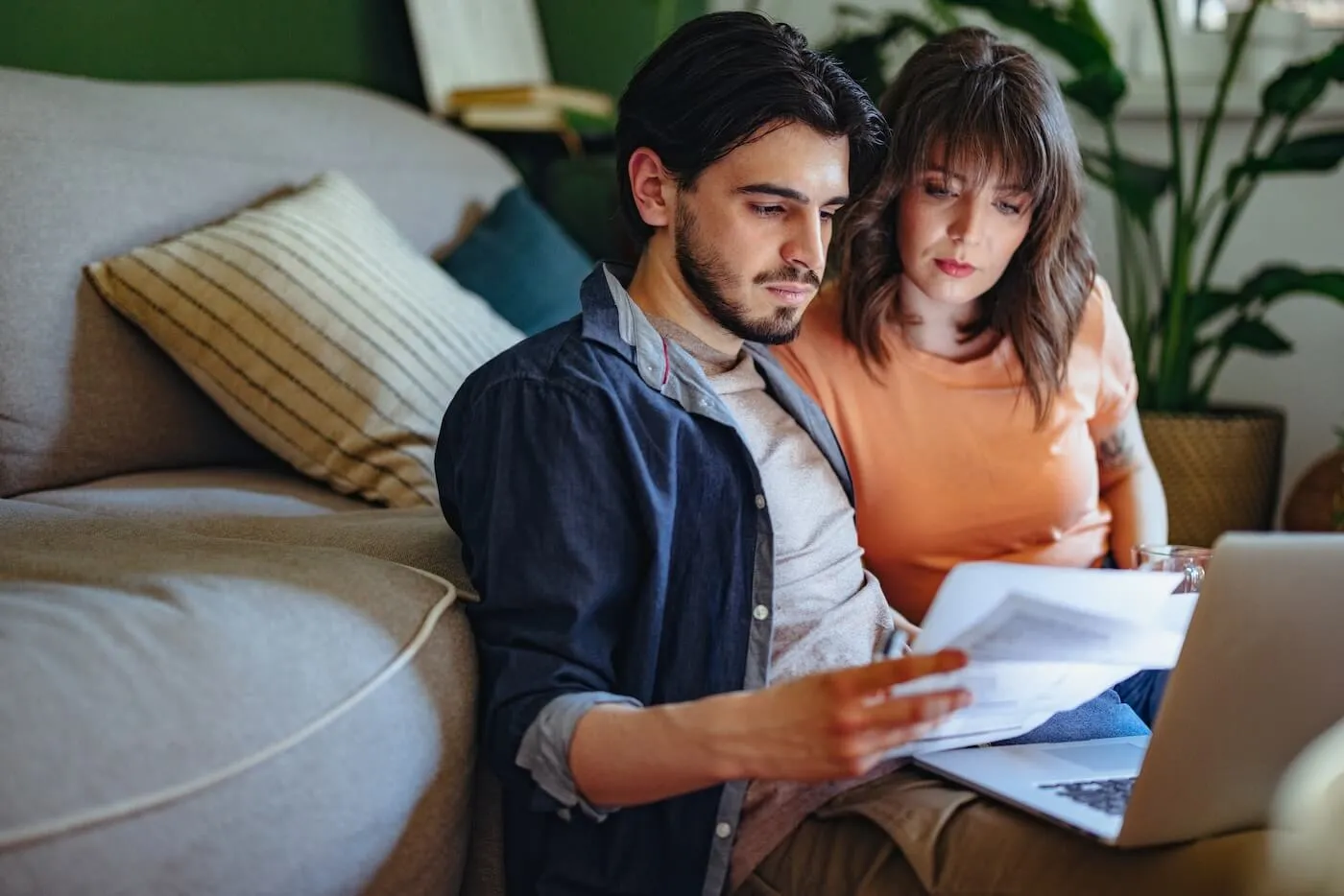 Focused couple sitting on the floor and leaning against a sofa while reviewing printed documents, with an open laptop on one person’s lap