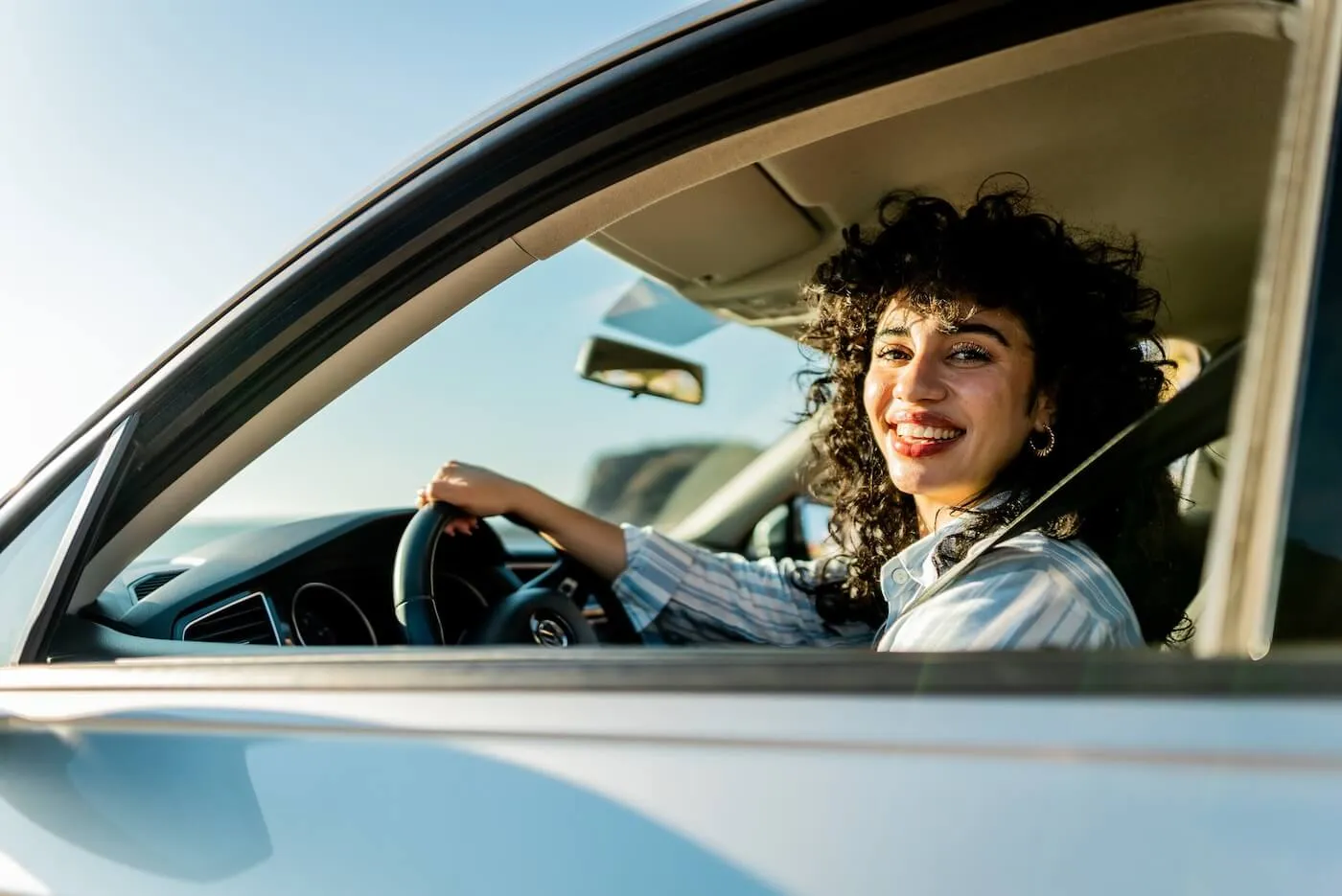 Smiling young woman driving a car along a coastal road, viewed from the driver’s side window.