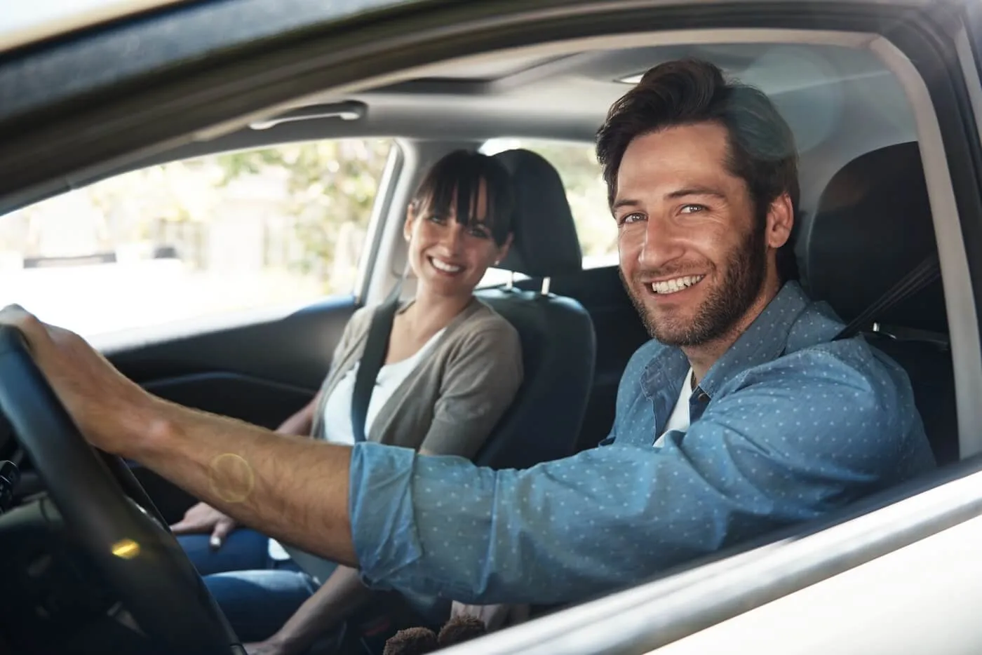 Happy couple sitting inside a car, with the man holding the steering wheel and the woman seated in the front passenger seat, both wearing seat belts