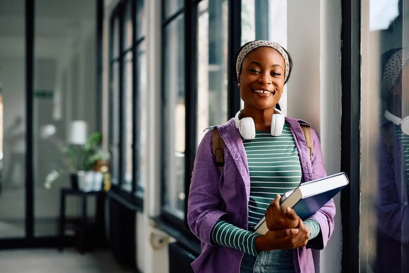 Smiling young woman with a backpack stands near tall windows in a modern hallway, holding a book and wearing headphones around the neck, with indoor plants visible in the background.