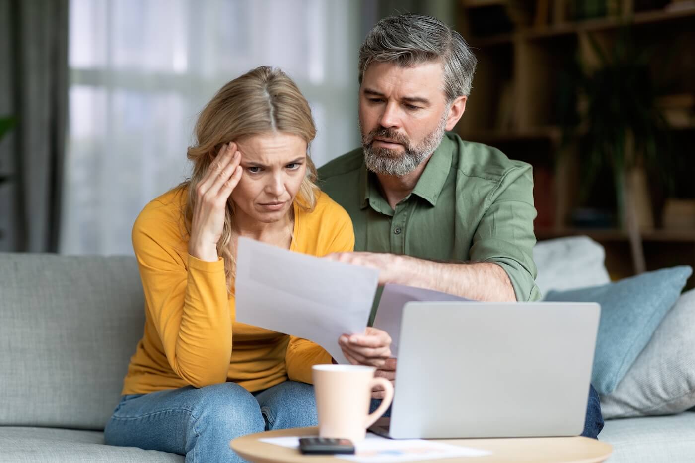 Concerned mature couple reviewing a printed document in the living room, with a laptop and a mug beside them on a coffee table