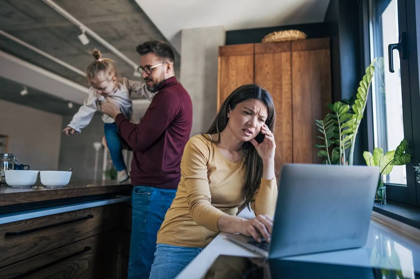 Concerned woman using a laptop at a kitchen island, while a man stands behind holding a small child; wooden cabinets, a countertop with bowls, indoor plants, and a large window are visible in the background.