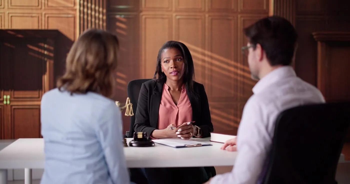 Legal professional seated at a desk meeting with two clients in a wood-paneled office, with legal books, paperwork, and a gavel visible on the desk during a consultation.
