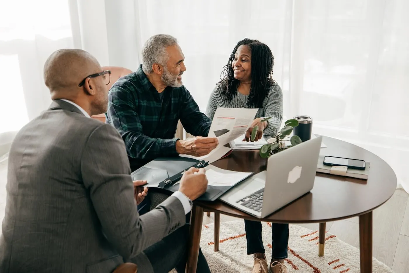 Two clients seated at a round table reviewing documents with a financial professional, with a laptop, paperwork, folders, and a smartphone visible during a consultation in a bright home office setting.