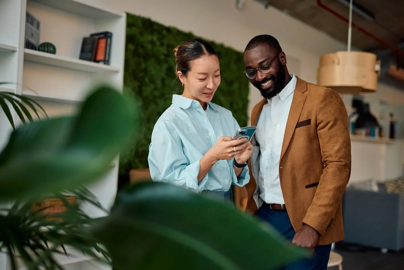 Two coworkers standing in a modern office, looking at information on a smartphone together, with plants, shelving, and a green wall feature visible in the background.