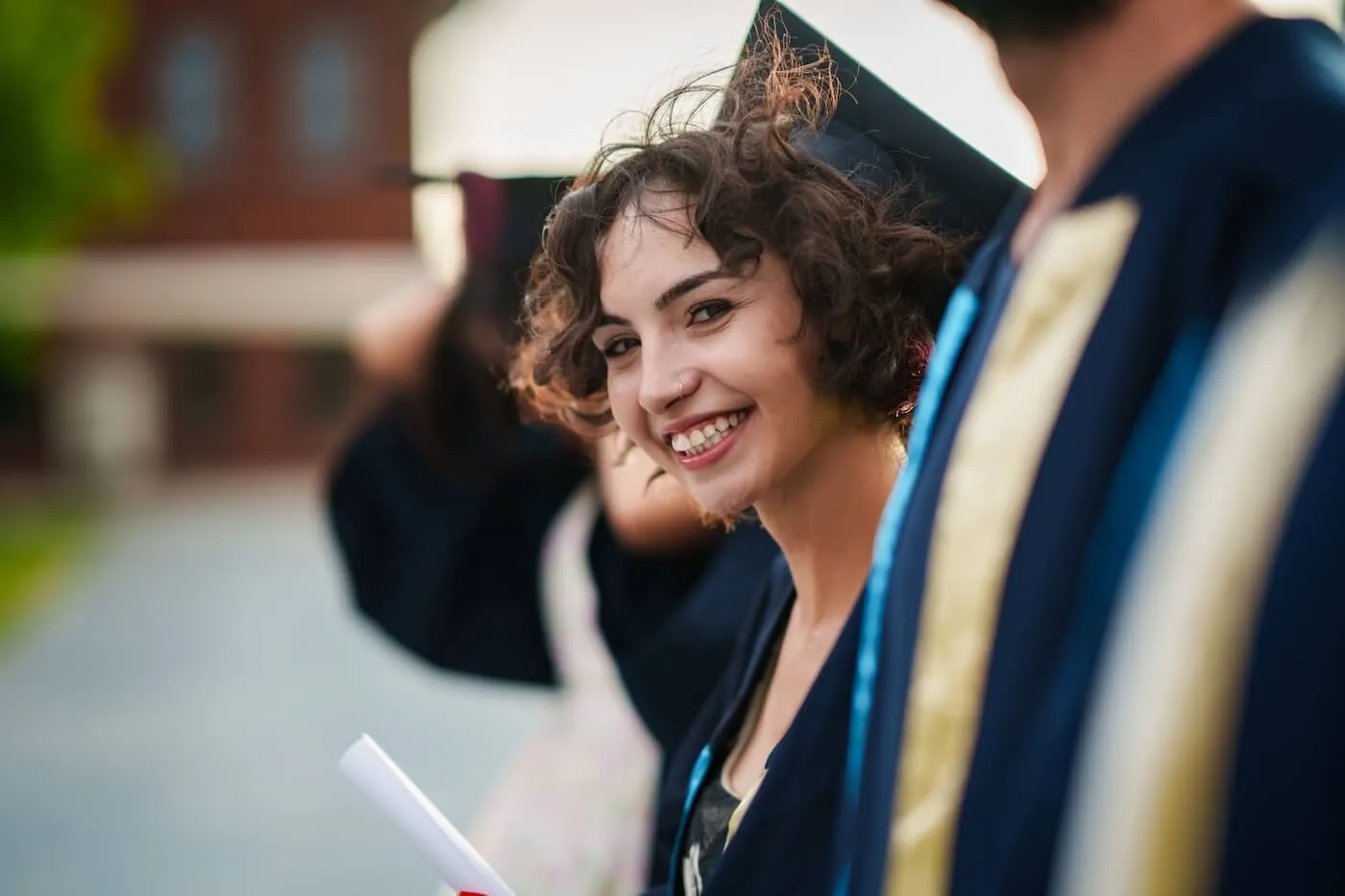 Smiling female graduate wearing a cap and gown and holding a diploma during an outdoor graduation ceremony, with other graduates and a campus building visible in the background.