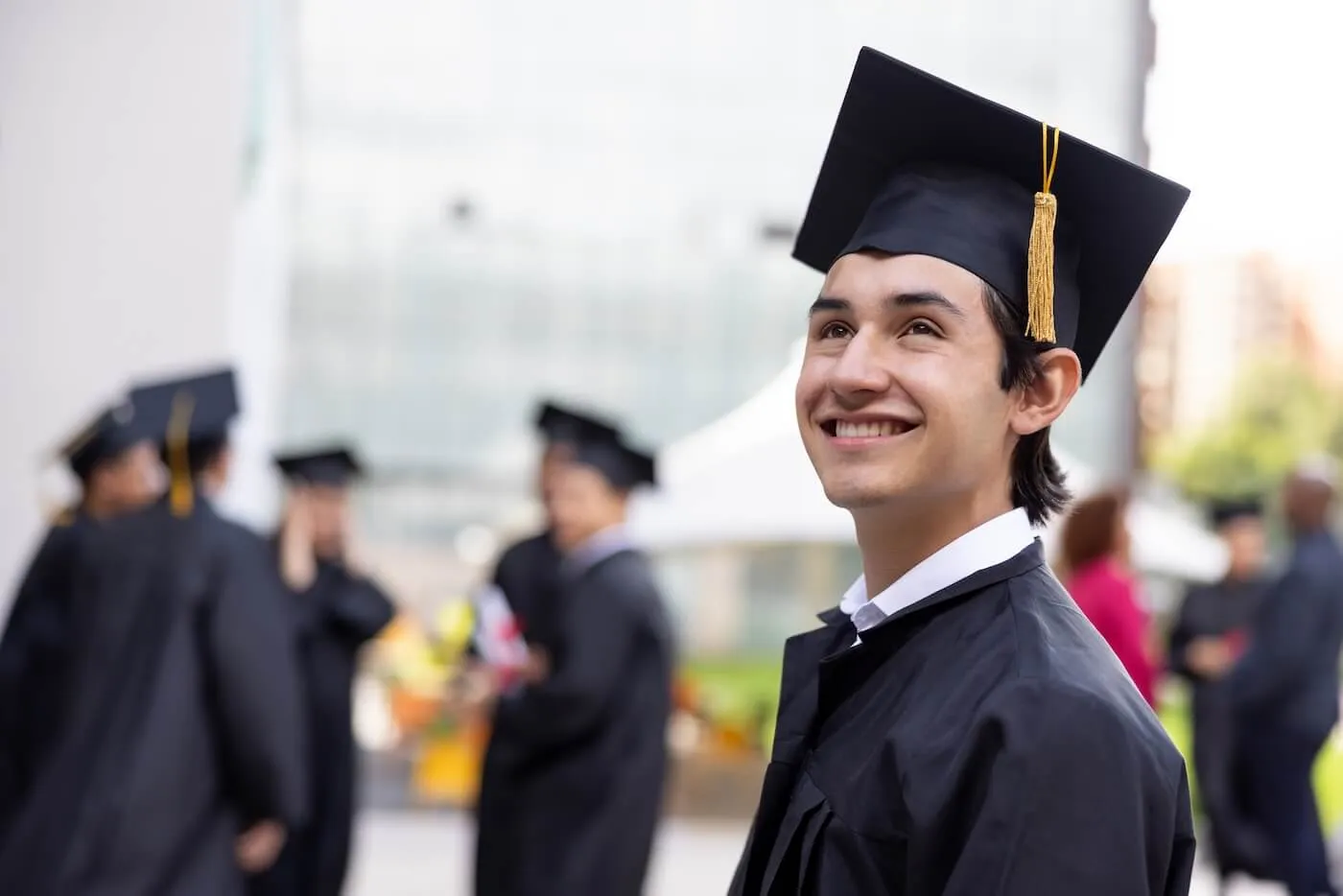 Smiling young man wearing a graduation cap and gown at an outdoor commencement ceremony, standing among other graduates with a campus building and attendees visible in the background.