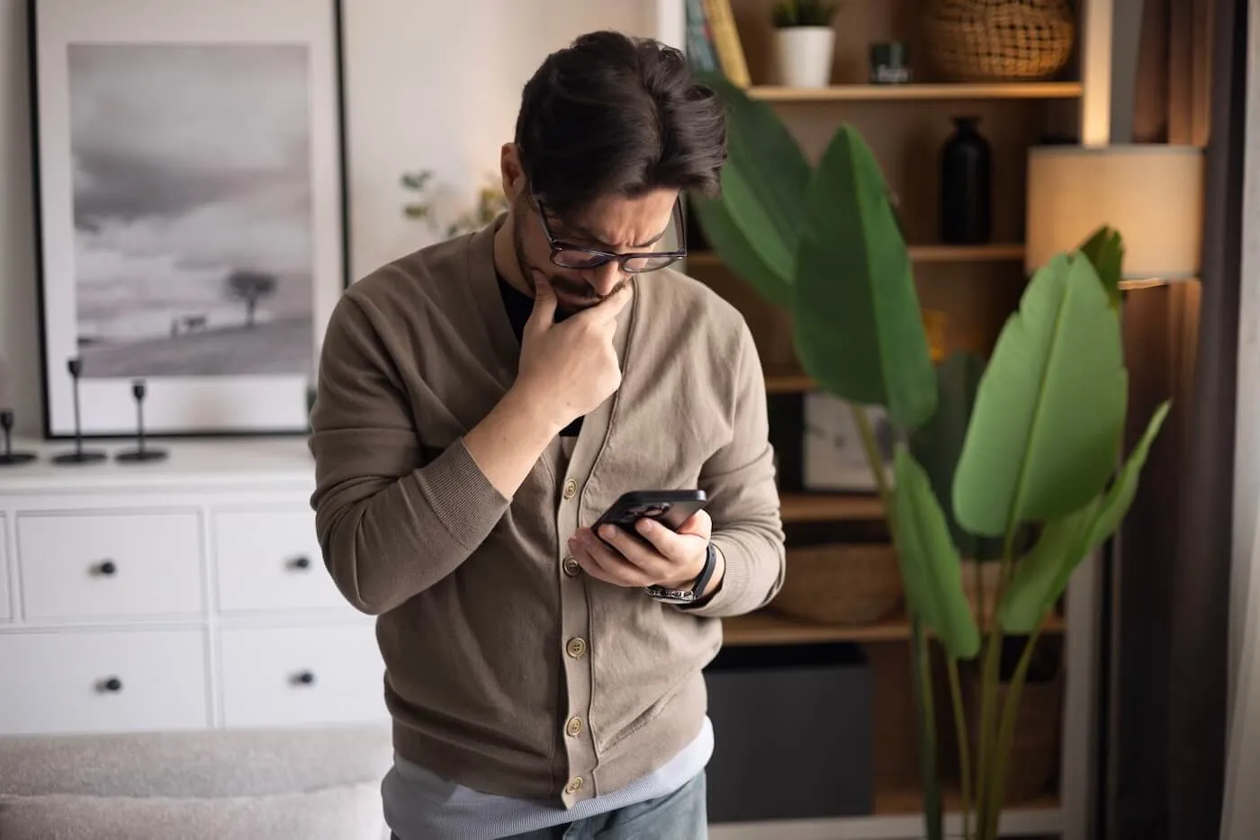 Concerned man standing in a living room and looking at a smartphone while resting a hand on the chin. Bookshelves, framed wall art, indoor plants, and a sofa are visible in the background.