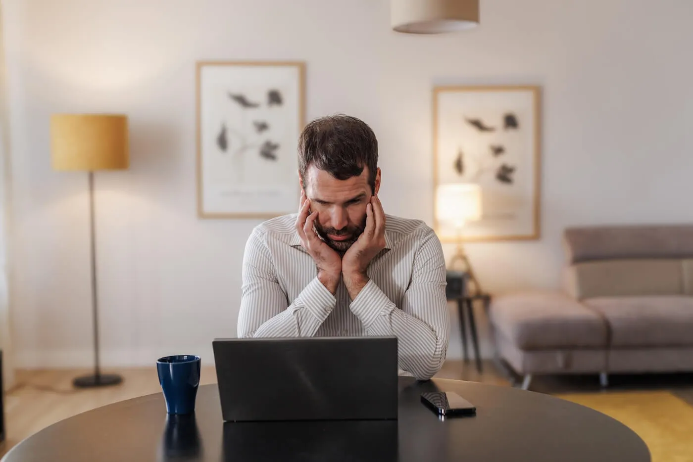 Concerned man seated at a round table in a living room, looking at a laptop computer. A coffee mug and smartphone rest on the table, while a sofa, floor lamp, and framed wall art are visible in the background.