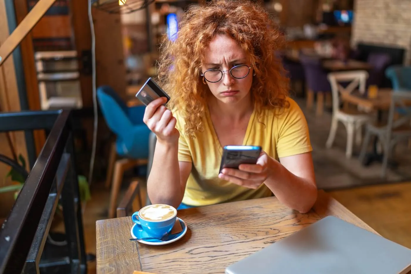 Concerned young woman seated at a café table holding a credit card while looking at a smartphone. A cup of coffee and a closed laptop sit on the wooden table, with café seating and décor visible in the background.