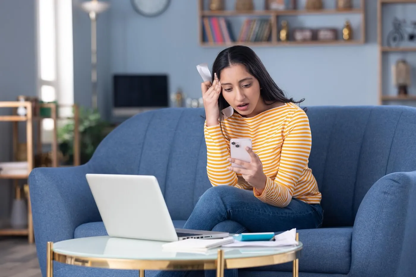 Concerned young woman seated on a blue sofa in a living room, looking at a smartphone while holding a receipt or bill. An open laptop, paperwork, and a notebook sit on a coffee table in front of the sofa, with bookshelves and home furnishings visible in the background.