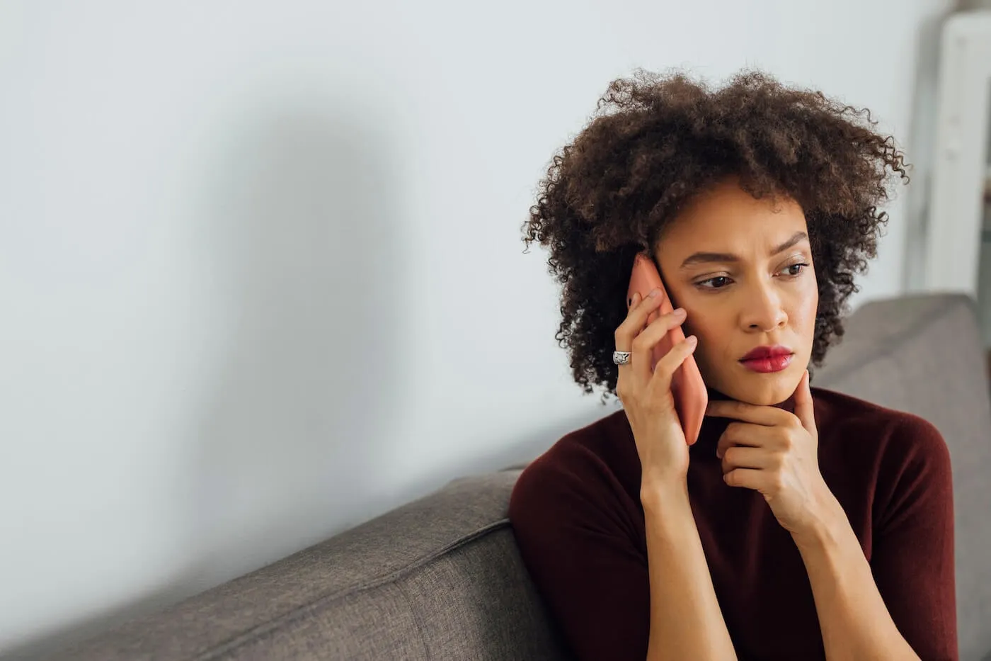 Concerned woman seated on a sofa in a home setting, holding a smartphone to one ear while looking off to the side. The image features a neutral-colored wall, soft natural lighting, and minimalist furnishings.