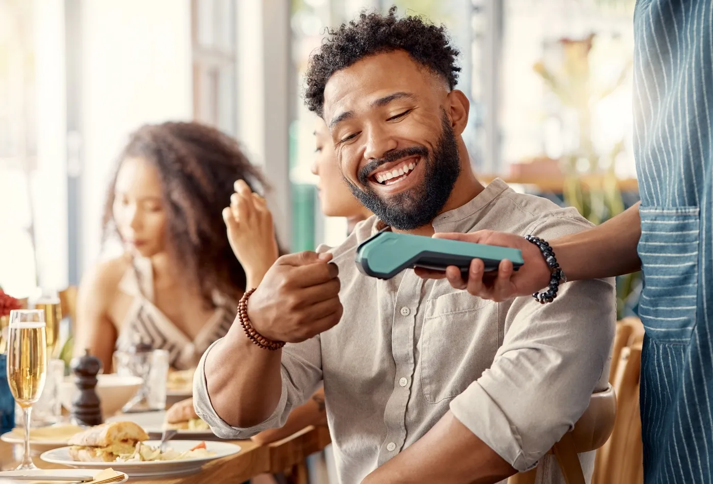 Smiling man seated at a restaurant table making a contactless card payment with a payment terminal held by a server, with plated food, beverages, and other diners visible in the background.