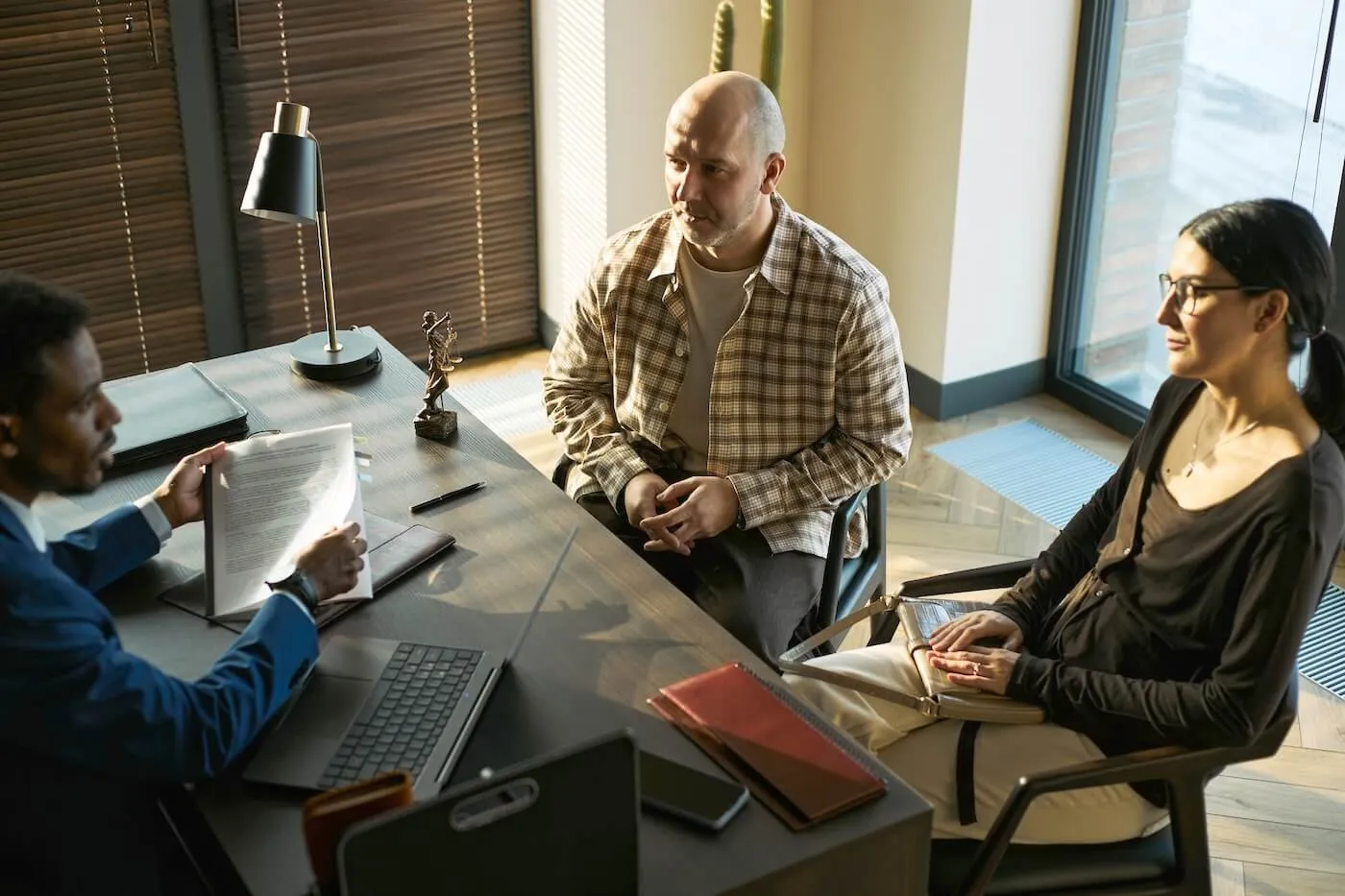 A couple seated across from a professional at a desk in a modern office, reviewing documents during a consultation, with a laptop, paperwork, and office furnishings visible in the meeting room.