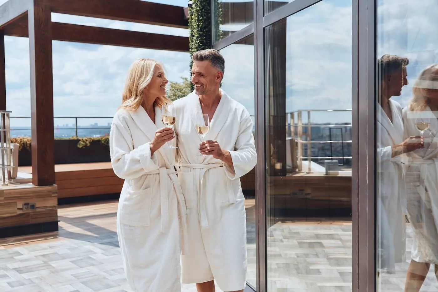 Two adults wearing white spa robes standing on an outdoor terrace and holding glasses of white wine, with a modern resort setting, glass walls, wooden architectural features, and a city skyline visible in the distance.