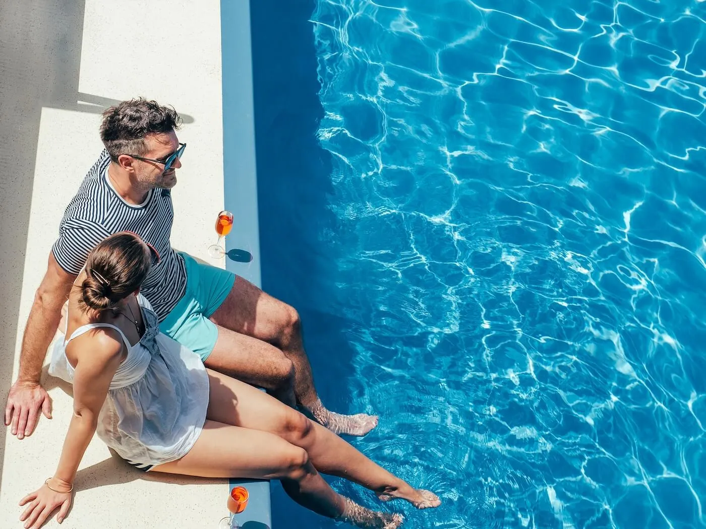 Relaxed couple sitting on the edge of a swimming pool with their feet in the water at a resort or vacation property. Drinks are placed beside them on the pool deck, and the bright blue water reflects sunlight.