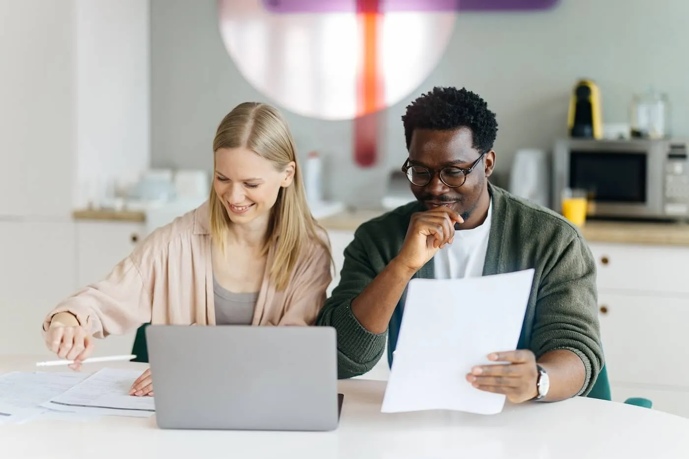 Happy couple sitting at a table reviewing financial documents and using a laptop in a home office or kitchen setting, with paperwork spread across the table.