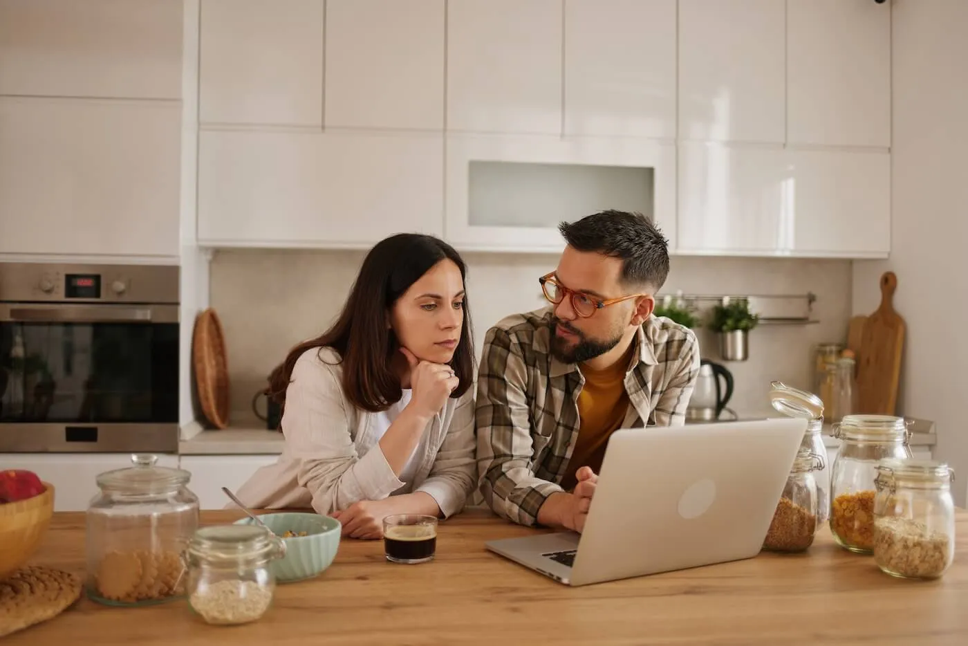 Focused couple sitting at a kitchen table reviewing information on a laptop, surrounded by food storage jars and household items in a modern home setting.