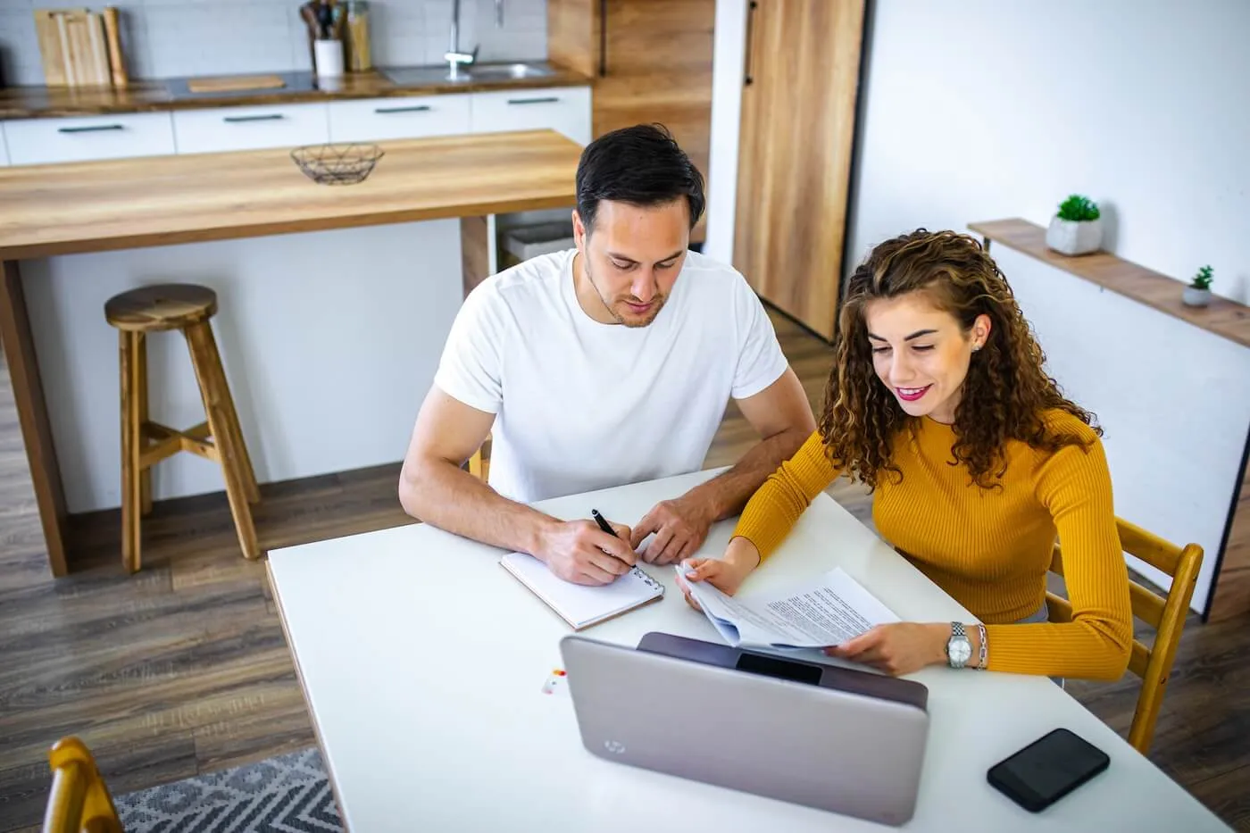 A couple seated at a table in a modern home, reviewing financial documents and taking notes beside an open laptop. A smartphone rests on the table, with a kitchen island, wood cabinetry, and contemporary furnishings visible in the background