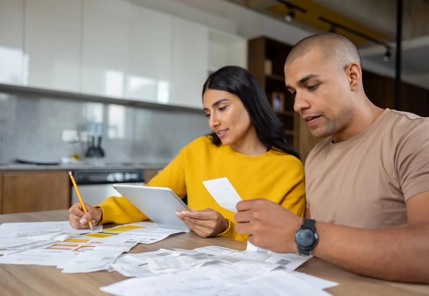 Focused couple sitting at a table covered with receipts and financial documents, using a tablet and reviewing paperwork in a home kitchen setting.
